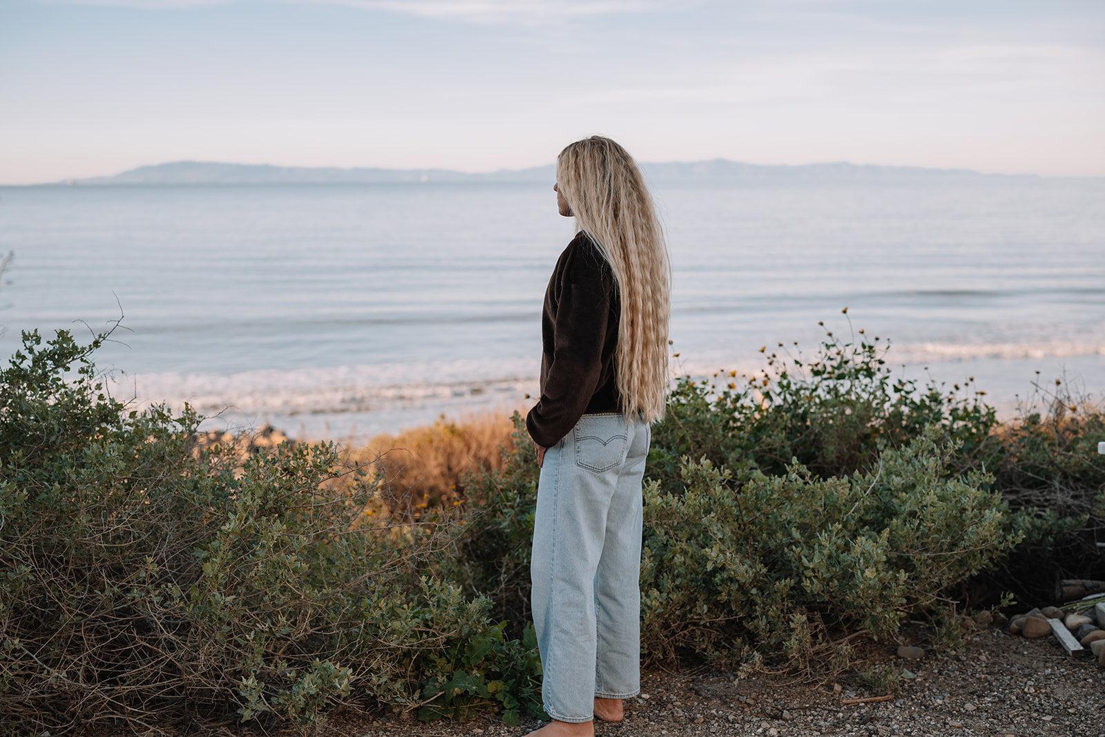 Woman with long blonde hair wearing a dark brown fleece sweater and light wash jeans, overlooking the ocean.