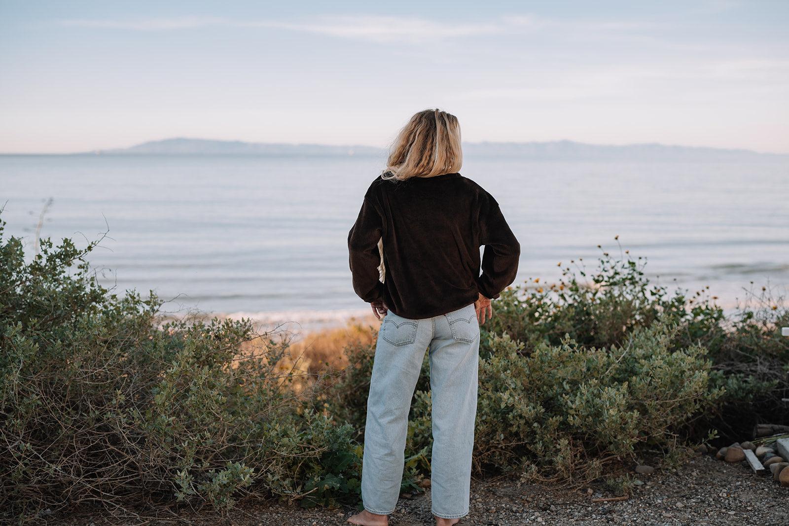 Blonde person in a dark brown Harborview Fleece Sweater and light wash jeans, facing the ocean.