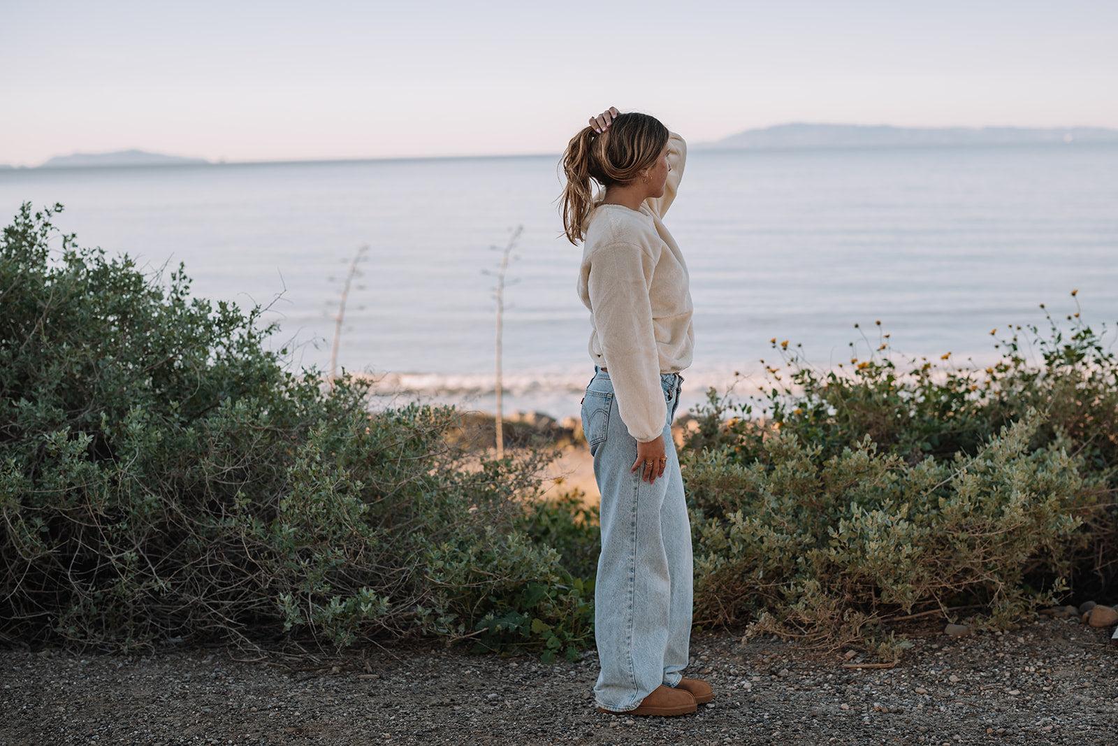 Woman in a cream Harborview Fleece Sweater and light wash jeans, standing by the ocean at sunset.