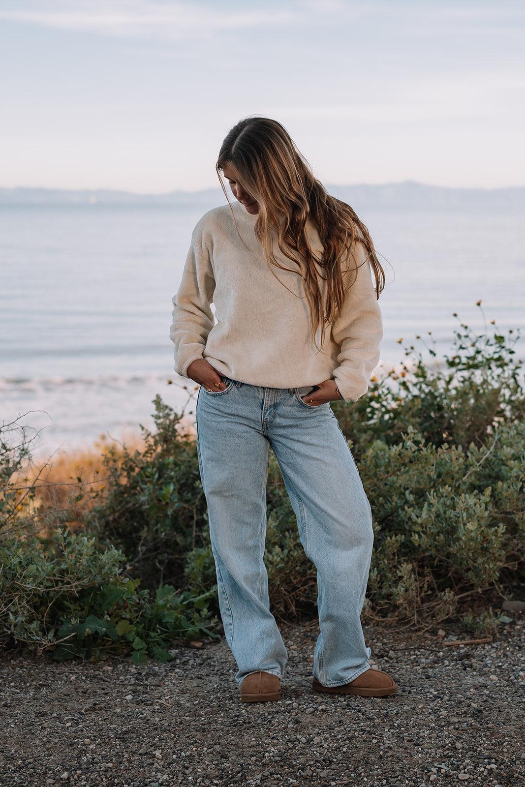 Woman in a cream Harborview Fleece Sweater and jeans, standing by the ocean.
