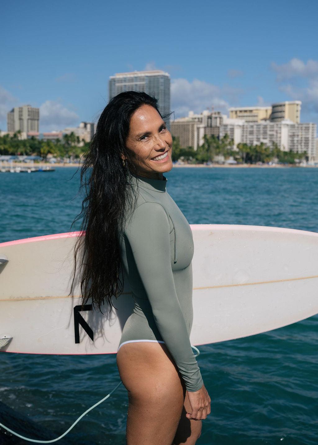 Smiling woman in a sage green long sleeve surf suit with a mock neck, standing by a surfboard and ocean.