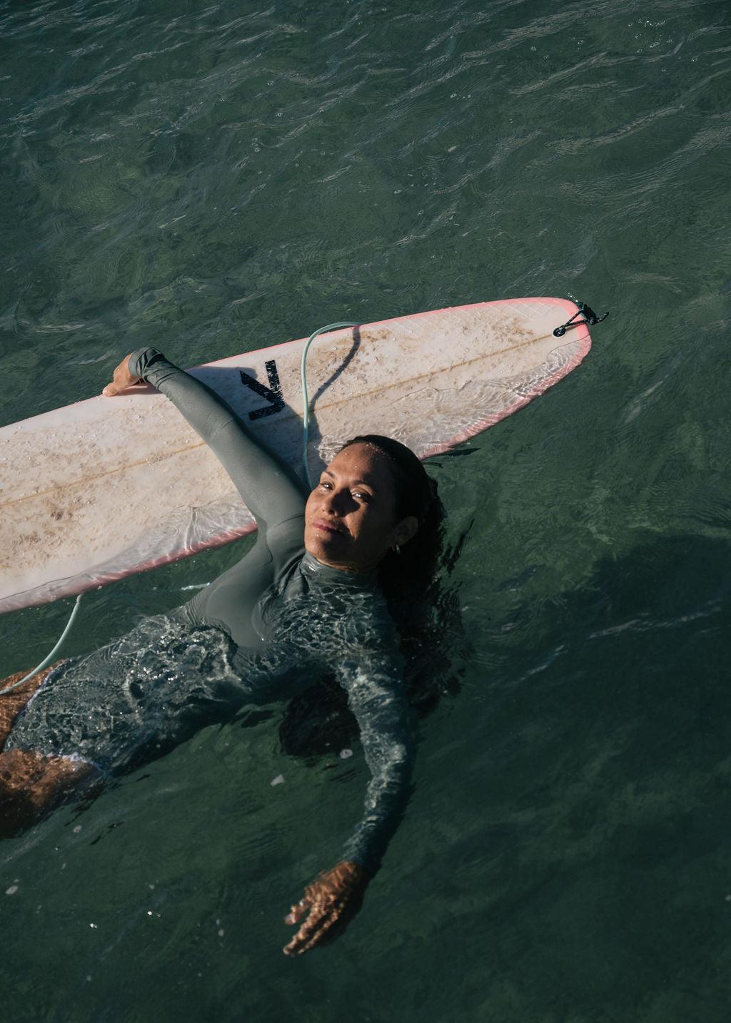 Woman in a gray long-sleeve Summer Shade surf suit floating in water with a surfboard.