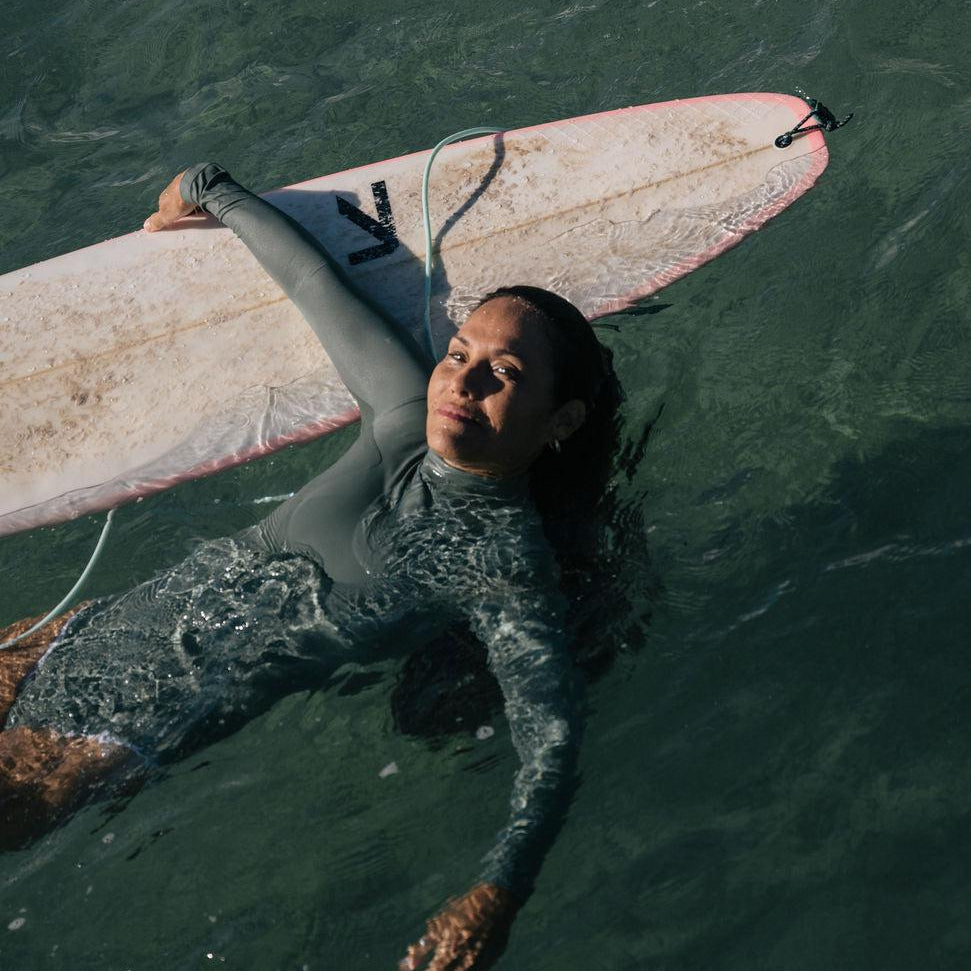 Woman in a gray long-sleeve Summer Shade surf suit floating in water with a surfboard.