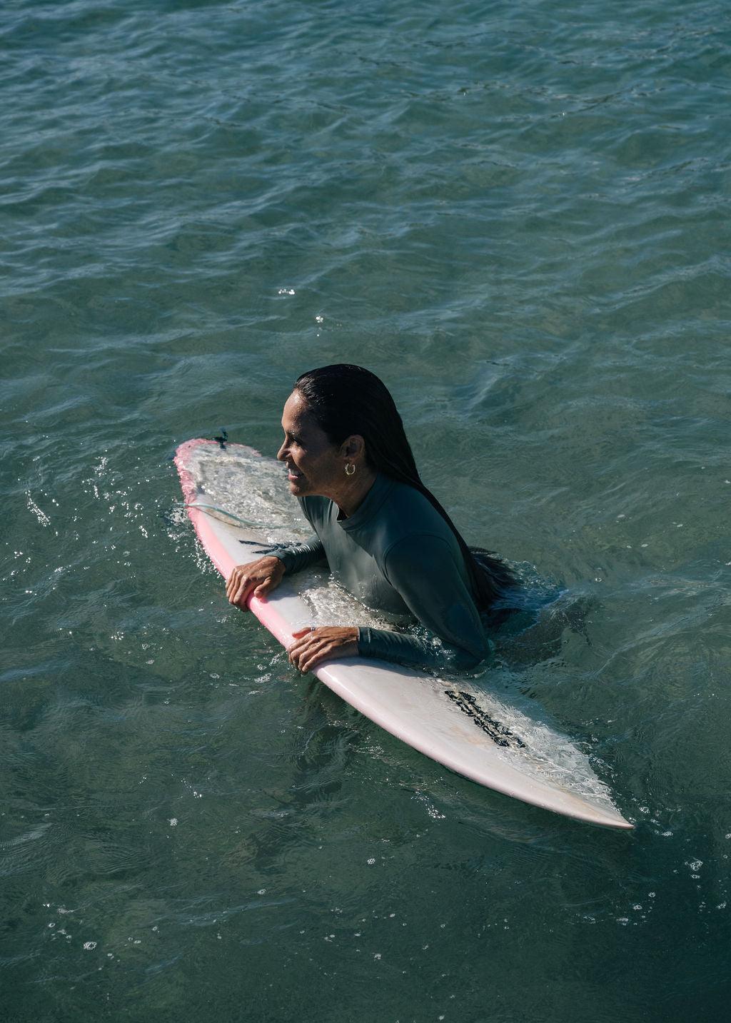 Woman in a dark teal long sleeve surf suit with a mock neck, holding a pink surfboard in clear water.